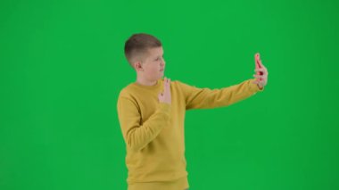 Modern çocuk okulu ve eğlence zamanı reklam konsepti. Krom anahtar yeşil ekrandaki çocuk portresi. Akıllı telefondan selfie çeken, gülen yüz ifadesi takınan okul çocuğu. Orta çekim.