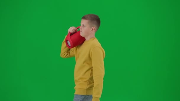 現代の子供の学校とレジャー時間の広告の概念 クロマキーグリーンスクリーンの少年肖像画 赤い音楽スピーカーを抱えて 音楽を聴くカジュアルなウォーキングでボーイ ミドルショット — ストック動画