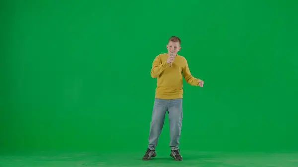 School kids and leisure time creative concept. Portrait of kid boy on chroma key green screen. Schoolboy in jeans holding holding karaoke microphone and singing song. Full body front shot.