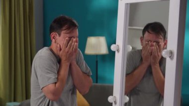 Yorgun bir adam aynadaki yansımasına bakıyor, oturma odasında dikiliyor. Duyarsızlık, iç çatışma, psikolojik sorunlar