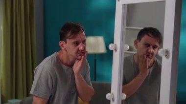 Yorgun bir adam aynadaki yansımasına bakıyor, oturma odasında dikiliyor. Duyarsızlık, iç çatışma, psikolojik sorunlar