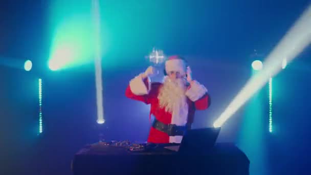 Djサンタクロースは クリスマスや新年のパーティーや企業イベントでナイトクラブでトラックを混合します サンタリスニング音楽 ヘッドフォン ラップトップ ミキサーコントローラープレーヤー ターンテーブルとしてのシニアディスクジョッキー — ストック動画