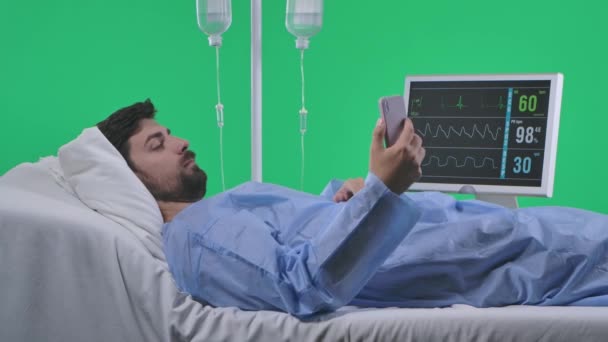 医療区 医療リハビリテーション広告コンセプト ドリップ付きのベッドで 心臓を呼吸して モニターをチェックする スマートフォンでセルフィーを取る男 クロマキーグリーンスクリーンに分離 — ストック動画