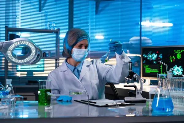 Female scientist sitting in a research lab. A female doctor in a white coat studying a sample of green liquid in a test tube. The concept of a bio or chemistry lab