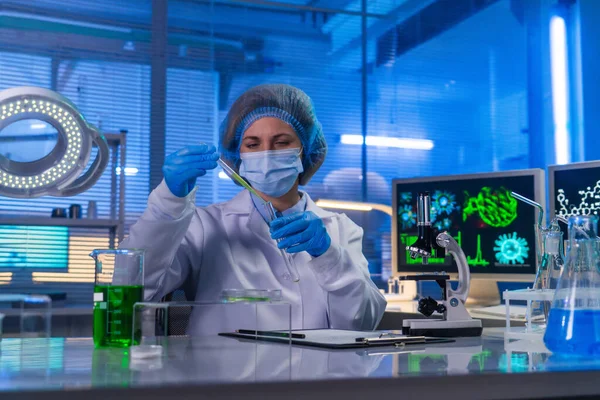 Female scientist sitting in a research lab. A female doctor in a white coat is pouring a sample of green liquid into a test tube using a pipette. Concept of a bio or chemistry lab