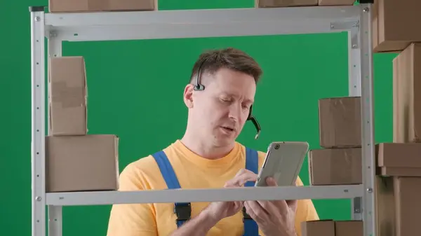 Framed on green background, chromakey. Depicted is an adult male wearing a work uniform. Demonstrates a storekeeper in a warehouse. He is wearing headphones and looking intently at a tablet. Medium.