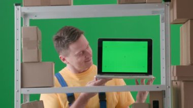 Yeşil arka plan kromatonuna göre çerçevelenmiş. İfade, iş üniforması giyen yetişkin bir erkek. Depodaki bir depocuyu gösteriyor. Kameraya bakan ve gülümseyen bir dizüstü bilgisayar tutuyor..