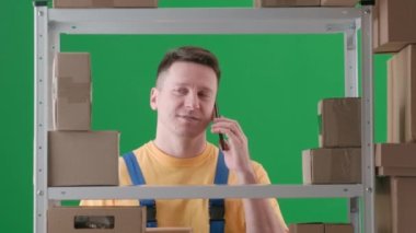 Yeşil arka planda çerçevelenmiş, kromakey. İş üniformalı yetişkin bir adam. Depoda bir depocuyu gösteriyor. Telefonda konuşuyor, bir şeyi tartışıyor, özenli, tatmin olmuş..