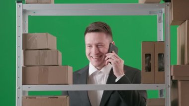 Çerçevede yeşil bir arka plan var. Takım elbiseli yetişkin bir adamı gösteriyor. Yöneticiyi gösteriyor, bir depodaki patronu. Telefonda konuşan kutulara bakar ve neşeyle gülümser. Orta çerçeve.