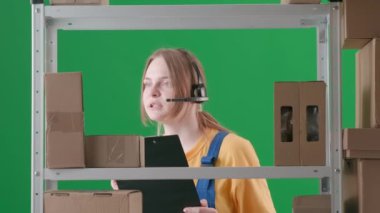 Yeşil arka planda çerçevelenmiş, kromakey. Üniforma ve kulaklık takan genç bir kadını gösteriyor. Depoda çalışan bir işçiyi, bir depocuyu gösteriyor. Bir tablet tutuyor ve bir şeye bakıyor..