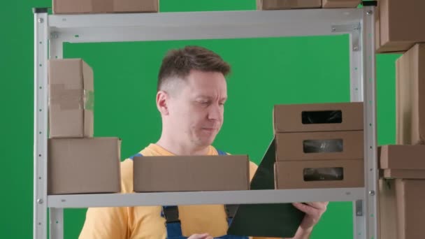 緑色の背景に 染色体 職場の制服を着ている男性です 倉庫に保管庫を設置しています 彼は箱を探し それを見つけ クリップボードにそれを書き込んで 葉を残しています — ストック動画