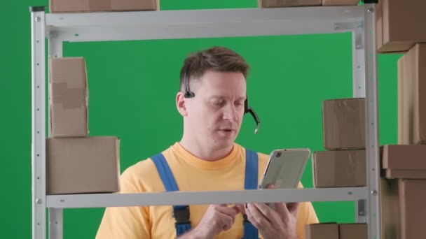 緑色の背景に 染色体 職場の制服を着ている成人男性です 倉庫に保管庫を設置する 彼はヘッドフォンを着て タブレットを見ている メディア — ストック動画
