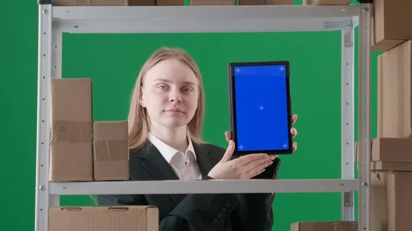 染色体の緑色の背景にあるフレーム スーツに若い女性がいる マネージャーは倉庫に展示されています 彼女はカメラを見て 微笑み 青い画面のタブレットを保持しています — ストック写真