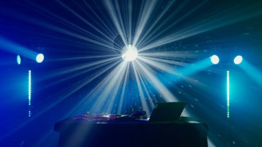 Bu büyüleyici görüntü, canlı bir ışık gösterisinin ortasında bir DJ kurulumu sergiliyor, yeşil ve mavi ışık demetleri DJ masasının üzerinde dramatik bir etki yaratıyor. Kulüp atmosferinin puslu havası