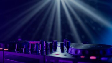 Bu görüntü, neon ışıklarla aydınlatılmış profesyonel bir DJ 'in ekipmanının yakın çekimini gösteriyor. Pikabın ve karıştırma tahtasının odak noktası, canlı renklerde parlayan düğmeler ve kaydıraklar