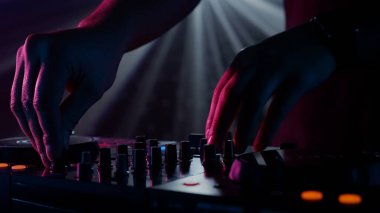 Fotoğraf, bir DJ 'in yetenekli ellerine ses mikserinin düğmelerini ayarlarken, ortam klubünün ışıklandırmasıyla yakınlaşıyor. Kırmızı ellerin canlı kontrastı yeşilin serin tonlarına karşı