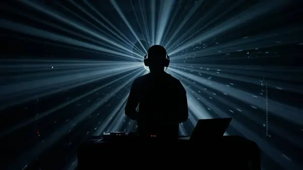 A silhouette of a male DJ with headphones stands against an illuminated backdrop of vibrant stage lights at a club event. The atmosphere is electric, with beams of blue and green light cutting through