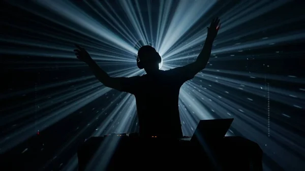A silhouette of a male DJ with headphones stands against an illuminated backdrop of vibrant stage lights at a club event. The atmosphere is electric, with beams of blue and green light cutting through