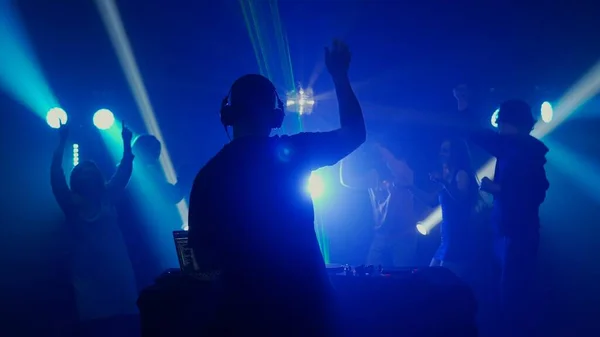 The image captures a DJ with arms raised, engaging the crowd at a live event. Backlit by the vibrant blue and green lights, the silhouette of the DJ and the audience creates an atmosphere of