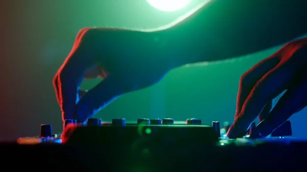 The photo zooms in on the skilled hands of a DJ adjusting the knobs of a sound mixer, with a backdrop of ambient club lighting. The vibrant contrast of red hands against the cool tones of green and