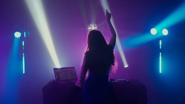 The image captures the exuberant moment of a female DJ raising her hand in joy, backlit by a spectrum of colorful stage lights at a nightclub. Shes wearing headphones and facing away from the camera