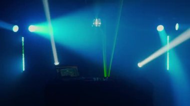 Bu büyüleyici video, canlı bir ışık gösterisinin ortasında, yeşil ve mavi ışık demetleriyle DJ masasının üzerinde dramatik bir etki yaratan bir DJ kurulumu sergiliyor. Kulüp atmosferinin puslu havası