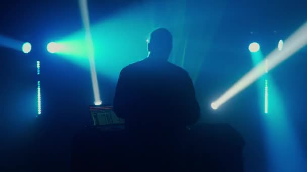 在俱乐部活动中 一位戴着耳机的Dj站在明亮的舞台灯光背景下 大气是电的 蓝光和绿光穿过 — 图库视频影像