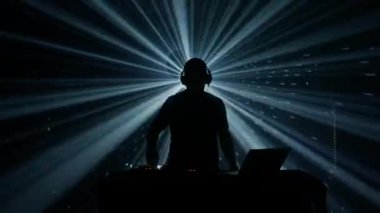 Kulaklıklı bir erkek DJ 'in silueti, bir kulüp etkinliğinde parlak sahne ışıkları karşısında duruyor. Atmosfer elektriklidir, mavi ve yeşil ışıklarla kesilir.