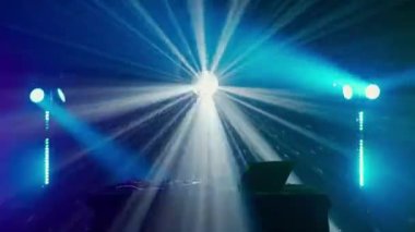 Bu büyüleyici video, canlı bir ışık gösterisinin ortasında, yeşil ve mavi ışık demetleriyle DJ masasının üzerinde dramatik bir etki yaratan bir DJ kurulumu sergiliyor. Kulüp atmosferinin puslu havası