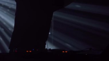 Kulaklıklı bir erkek DJ 'in silueti, bir kulüp etkinliğinde parlak sahne ışıkları karşısında duruyor. Atmosfer elektriklidir, mavi ve yeşil ışıklarla kesilir.