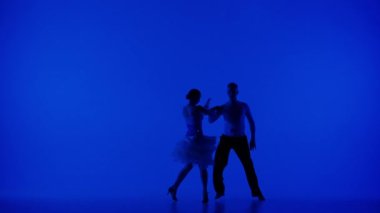 Zarif bir dans çifti, hareketlerini vurgulayan çarpıcı bir mavi arka plana ve beyaz etekli kadın dansçıların akışına karşı orta pozisyonda çekiliyor. Bej giysili adam.