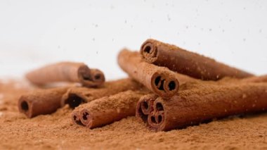 Aromatik baharatlar yaratıcı reklam konsepti. Yemek malzemelerinin stüdyo çekimlerini kapat. Kahverengi kurutulmuş tarçın çubukları beyaz zemine serilmiş toz ve tepeden düşen birkaç çubuk..
