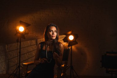 Film seti ve sahne arkası yaratıcı reklam konsepti. Çekici kadın oyuncu portresi. Siyah elbiseli esmer kadın. Saç stili ve incili. Yönetmen koltuğunda oturmuş kameraya bakıyor..