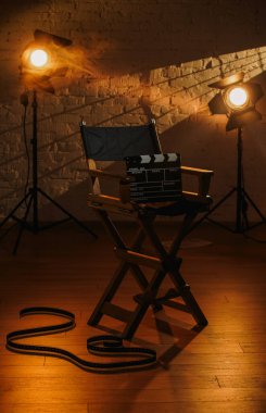 Film seti ve sahne arkası yaratıcı reklam konsepti. Sinema sehpasının yakın plan çekimi yönetmen koltuğunda, projektörler arka planda, sıcak ışık, film makarası yerde..