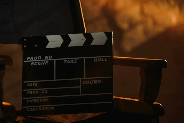 Movie set and backstage creative advertisement concept. Close up view of black clapperboard standing on directors chair, warm light from projectors on the wall at the background.
