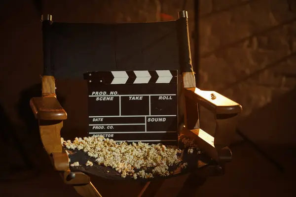 Movie set and backstage creative advertisement concept. Close up shot cinema clapperboard standing on directors chair popcorn scattered on chair, rays of light from the spotlights at the background.