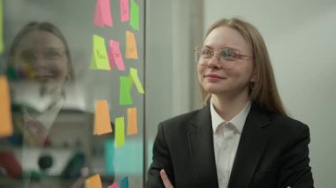 Modern iş dünyasının yaratıcı konseptinde beyin fırtınası ve fikirler. Ofis planlama projesindeki şirket liderinin portresi. Cam duvarın yanındaki iş kadını yapışkan notlarla dolu, gülen yüzlü..