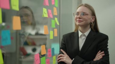 Modern iş dünyasının yaratıcı konseptinde beyin fırtınası ve fikirler. Ofis planlama projesindeki şirket liderinin portresi. Cam duvarın yanındaki iş kadını yapışkan notlarla dolu, gülen yüzlü..