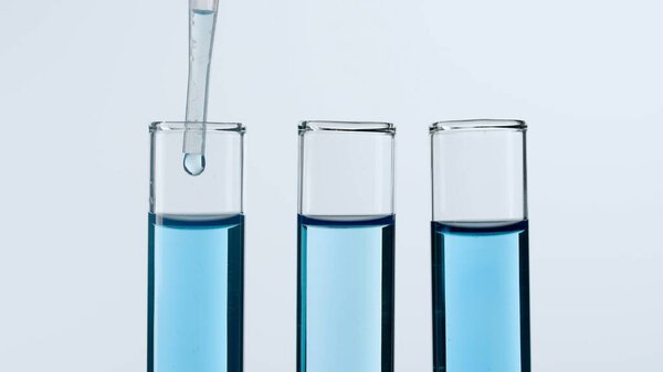 Three glass test tubes on a white background. Two test tubes are filled with blue liquid, a blue substance is dripping into one of them from a pipette. Concept of medicine, biochemical research. Close