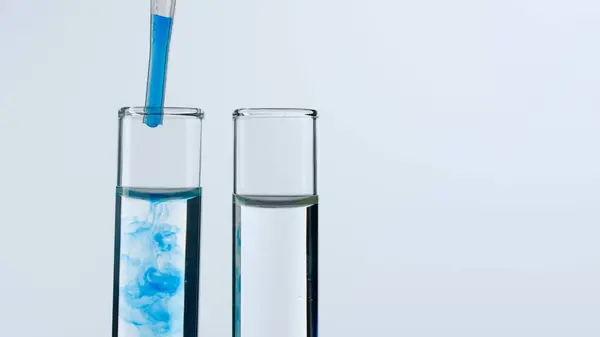 Two glass test tubes on a white background. Test tubes are filled with transparent liquid, a blue substance is dripping into one of them from a pipette. Concept of medicine, biochemical research