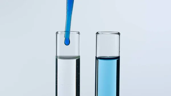 Two glass test tubes on a white background. Test tubes are filled with liquid, a blue substance is dripping into one of them from a pipette. Concept of medicine, biochemical research. Close-up