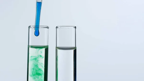 Two glass test tubes on a white background are filled with a transparent liquid. A blue substance drops into one of the test tubes from a pipette and dissolves in green color. Close up.