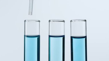 Bilim ve biyoteknoloji kavramı. Beyaz arka planda laboratuvar camlarının yakın plan görüntüsü. Mavi sıvıyla dolu test tüplerinin stüdyo çekimi, bilim adamı tüpün içine pipette sıvısı ekliyor. Yavaş çekim