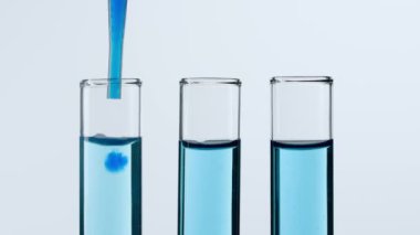 Bilim ve biyoteknoloji kavramı. Beyaz arka planda cam bardakları kapatın. Renkli sıvılarla dolu test tüplerinin stüdyo çekimi, bilim adamı tüpün içine mavi mürekkep sıvısı ekledi. Yavaş çekim.