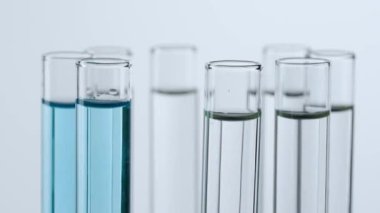 Beyaz arka plandaki laboratuvar camlarını kapatın. Platformda sıvı dönen test tüpleri. Test tüplerine şeffaf sıvı damlayan mavi madde damlaları. Bilim yaratıcı konsept.