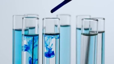 Beyaz arka plandaki laboratuvar camlarını kapatın. Platformda sıvı dönen test tüpleri. Test tüplerine şeffaf sıvı damlayan mavi madde damlaları. Bilim yaratıcı konsept.