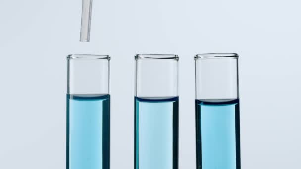 科学とバイオテクノロジーのコンセプト ホワイトバックのラボ用ガラス製品のクローズアップショット 青い液体で満たされた試験管のスタジオショット チューブからの科学者のピペット液体 スローモーション — ストック動画