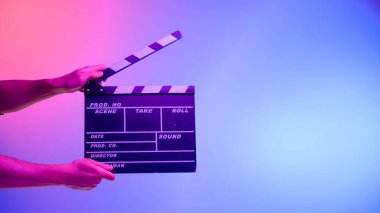 El, siyah bir el çırpma tahtası ya da film tahtası tutuyor. Mavi arka planda, pembe neon ışıkla aydınlatılmış dairesel bir ışık var. Sinema endüstrisi, eğlence