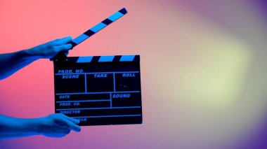 El, siyah alkış tahtasını ya da pembe arka planda mavi neon ışıkla aydınlatılmış bir film tahtasını tutuyor. Sinema endüstrisi, eğlence