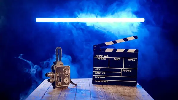 A clapper board and vintage video camera on a wooden table enveloped in smoke. Cinematography equipment in studio on black background in blue neon light